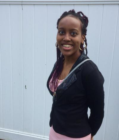 A black woman with braids and gold hoop earrings smiling at the camera in a pink dress and black sweater.