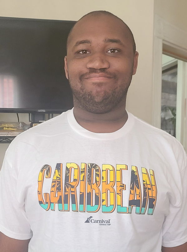 A dark skinned man in a white tshirt, with the words "Caribbean" on it, smiling at the camera