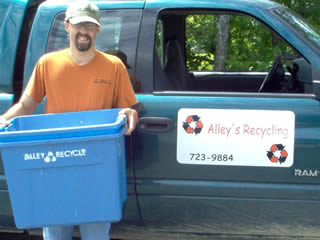 Holding a blue recycling bin, Corey smiles in front of a truck with 'Alley’s Recycling' and the business’s phone number marked on its passenger side door.