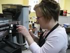 Emilea, wearing a black apron, stands in front of a machine in the coffee shop, with her left hand using the machine to fill the cup held in her right hand.