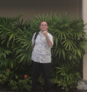 Ian stands in front of some bushes and flowers, with a backpack on his right shoulder, facing the camera and making a shaka sign with his left hand.