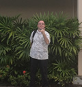 Ian stands in front of some bushes and flowers, with a backpack on his right shoulder, facing the camera and making a shaka sign with his left hand.