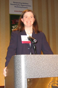 Mia stands at a podium, with a microphone in front of her, hands grasping the sides of the podium, and smiling