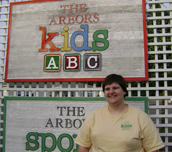 Taking steps from part-time classroom assistant toward full-time childcare professional Nicole smiles in front of a colorful sign for The Arbors Kids.