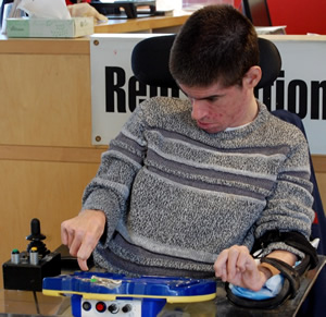 Cody looks down at his voice output machine, with his hand over one of the buttons.