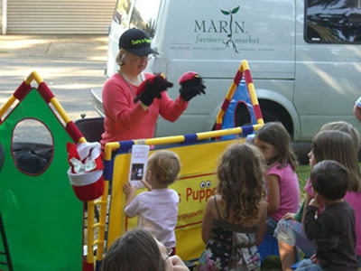 Dusty, who stands in front of a colorful makeshift barrier, wearing a puppet on each hand, is watched by a group of children.