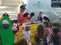 Dusty, who stands in front of a colorful makeshift barrier, wearing a puppet on each hand, is watched by a group of children.