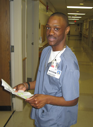 Emmitt, wearing scrubs and an ID badge, holds a piece of paper with both hands and looks towards the camera on his left.