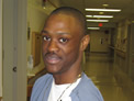 Emmitt, wearing scrubs and an ID badge, holds a piece of paper with both hands and looks towards the camera on his left.