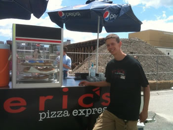 Eric stands in front of the 'Eric's Pizza Express' food cart with his right arm on the counter, wearing a T-shirt with a logo to match the front of the food cart.