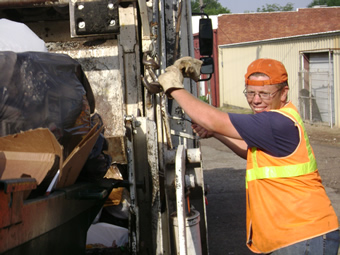 Dressed in orange vest and backwards orange hat, Frankie stands with both gloved hands holding onto the side of a truck and smiles towards the camera.
