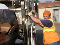 Dressed in orange vest and backwards orange hat, Frankie stands with both gloved hands holding onto the side of a truck and smiles towards the camera