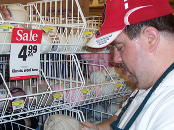 In this close-up photo, John looks down at a yarn product in his hand, as he stands in front of a sales display and sign, indicating that 'Classic Wool Yarn' is on sale.
