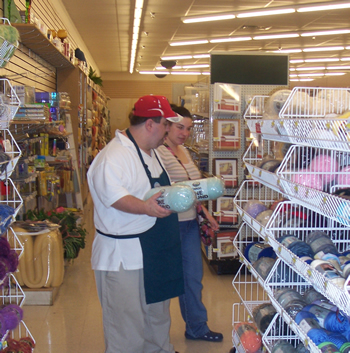 Dressed in his Jo-Ann’s Fabrics apron and a red hat, John stands in an aisle facing a sales display full of yarn and holds a yarn product in each hand, while a woman stands to his left looking at the yarn display.