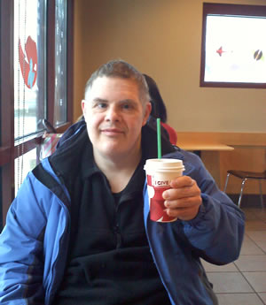 Seated in a cafe, Ken faces the camera and holds up a Starbucks beverage.