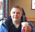 Seated in a cafe, Ken faces the camera and holds up a Starbucks beverage.