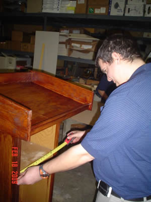 Matt holds measuring tape up to a piece of wooden furniture, in front of a wall of shelves stocked with boxes and other supplies