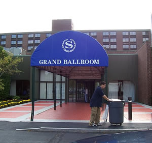 Outside in front of a blue awning for the Sheraton Grand Ballroom, Matt pushes a wheeled trash bin.