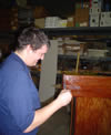 Matt uses a small tool on a piece of wooden furniture, in front of a wall of shelves stocked with boxes and other supplies