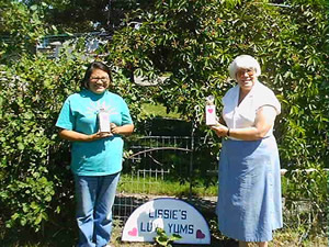 Standing in the sun in front of some trees and bushes, Melissa and her mother each hold a package of gourmet dog biscuits and stand on either side of a 'Lissie’s Luv Yums' sign.